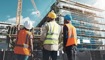 Three People In Hi-vis Vests And Hardhats Stand Just Outside A Black Construction Fence Around The Base Of A Large Commercial Building Being Built.