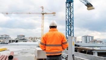 A Man In A Hard Hat And Orange Vest Examines Work At Commercial Building Under Construction.